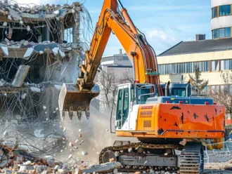 Ein Kettenbagger reißt ein Gebäude ab, während Beton und Bauschutt auf einer Baustelle herunterfallen.